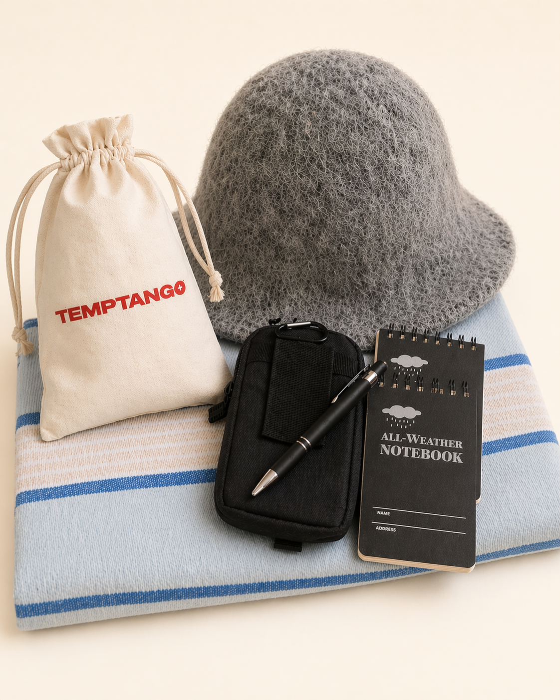Black waterproof bag, black waterproof notebook, pen, blue towel with a beige drawstring bag featuring 'Temptango' and a grey wool sauna hat. Sauna essential bundle